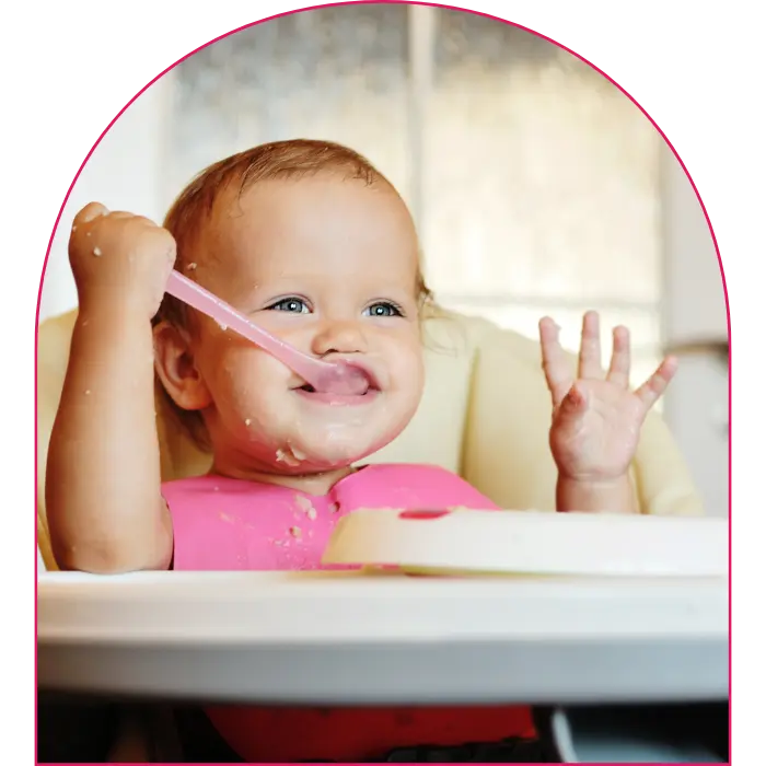 Little baby in high chair holding spoon and eating